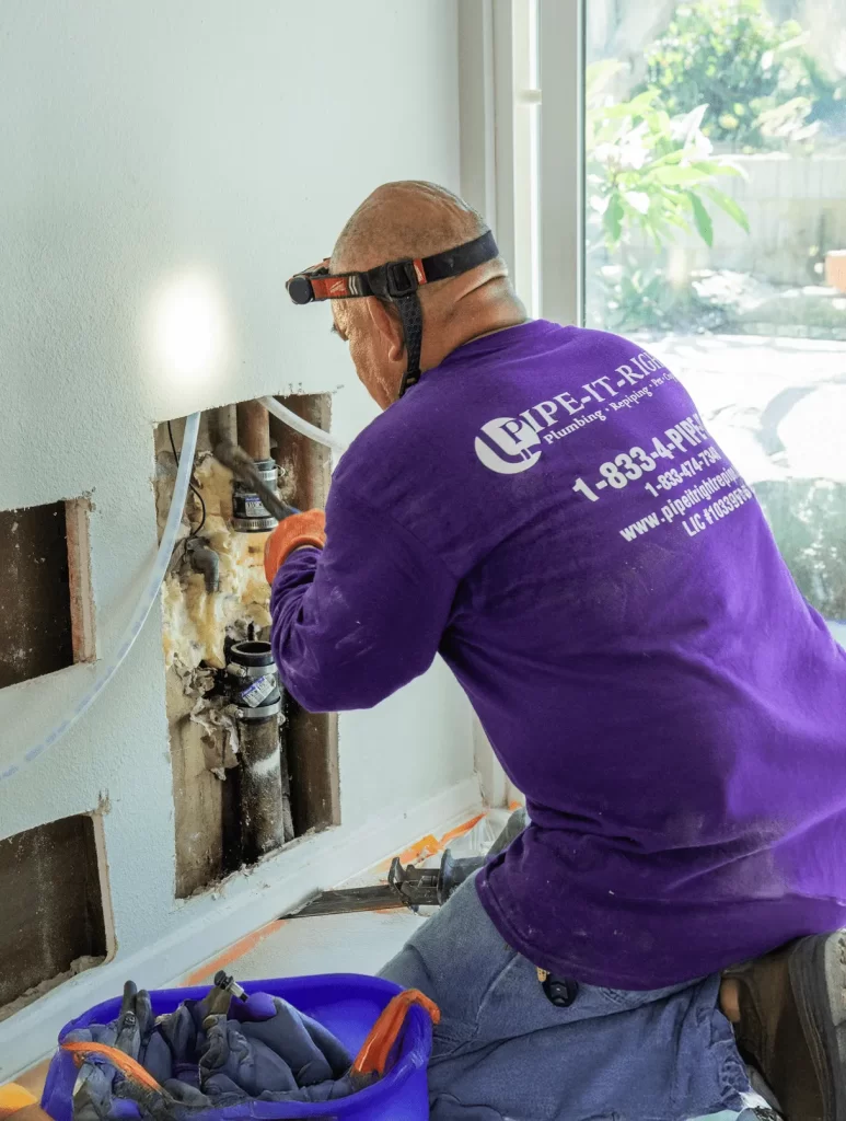 Plumber wearing a headlamp and purple shirt repairs water pipes inside a wall opening of a house.