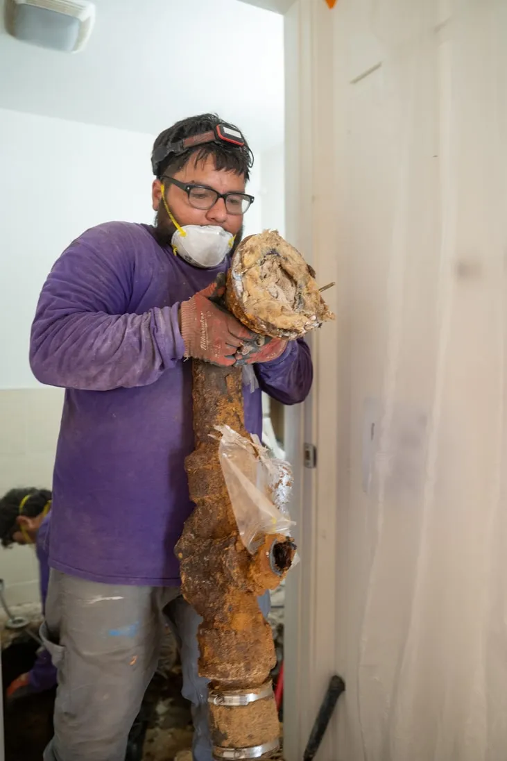 Plumber wearing protective gear removes a large, corroded pipe inside a building